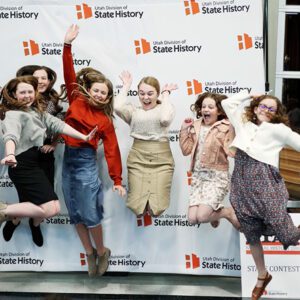 Teenage girls jump joyfully in front of a banner that reads Utah Division of State History