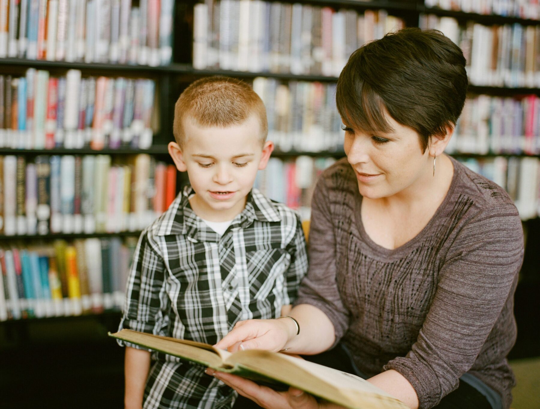 A woman with short brown hair reading a book to a young blonde boy in a library.