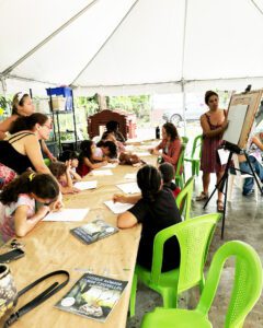 Women and children gather around a long table outside under a large tent. The kids are writing on pages while watching an instructor using a flipboard.
